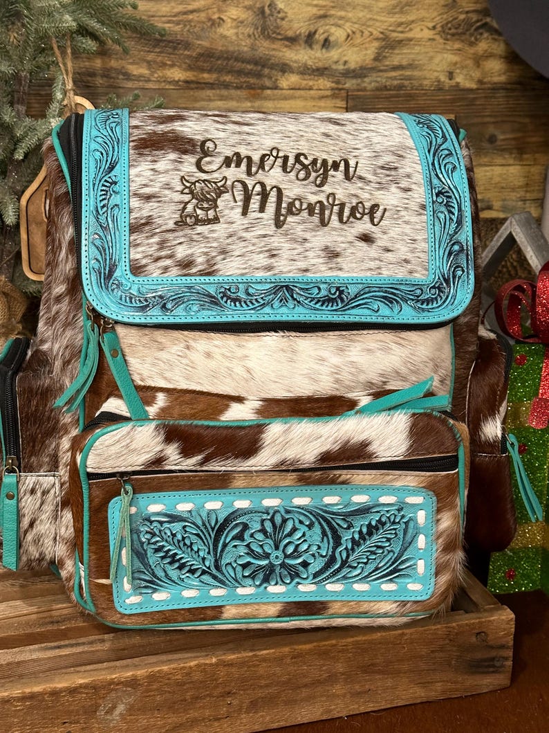 Op de afbeelding: Een bruin en wit koeienhuid rugzak met een turquoise lederen klep voorzien van reli&euml;f bloemmotieven, de naam Emersyn Monroe en een kleine koeienafbeelding. Het voorvak heeft een bijpassend turquoise lederen paneel met witte stiksels.