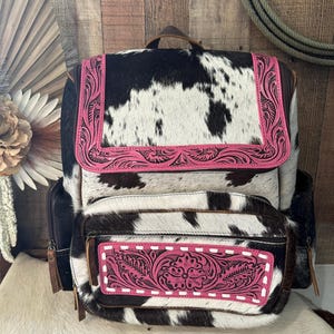 May include: A backpack featuring a black and white cowhide pattern with bright pink leather accents. The pink leather panels on the top flap and front pocket are decorated with intricate floral tooling and white stitching details.