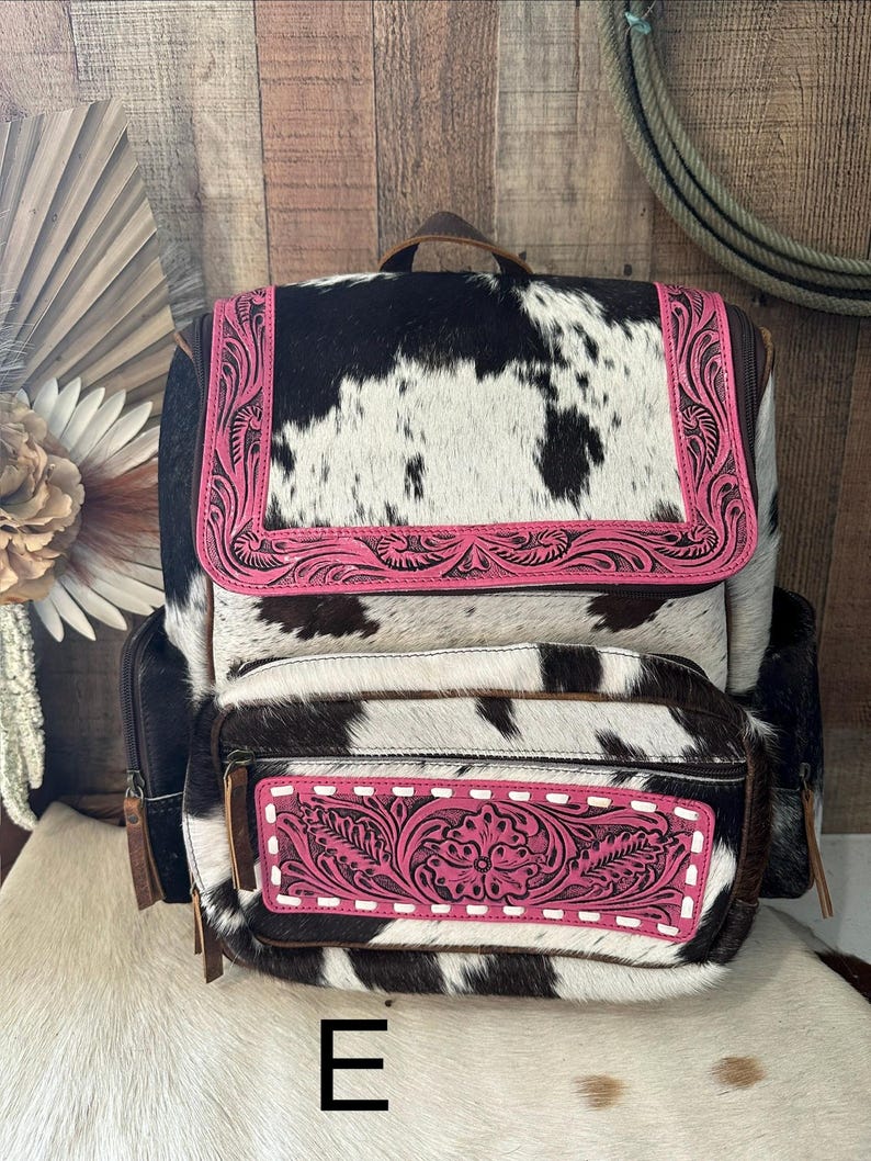 May include: A backpack featuring a black and white cowhide pattern with bright pink leather accents. The pink leather panels on the top flap and front pocket are decorated with intricate floral tooling and white stitching details.