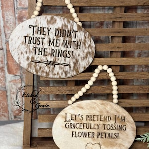 May include: Two oval wooden signs with a brown and white cowhide pattern. The top sign reads "THEY DIDN'T TRUST ME WITH THE RINGS!" with a barbed wire graphic. The bottom sign reads "LET'S PRETEND I'M GRACEFULLY TOSSING FLOWER PETALS!" with a heart.