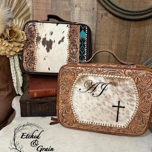 Peut inclure: Deux mallettes rectangulaires en cuir avec poignées supérieures. Les panneaux avant présentent des inserts en peau de vache blanche et brune entourés de motifs floraux gravés. L'une affiche les lettres AJ et une croix noire.