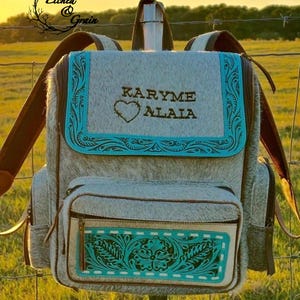 May include: A backpack made of grey and white cowhide featuring turquoise leather trim with embossed floral patterns. The front flap displays the text KARYME ALAIA above a heart outline. It includes a front zippered pocket and brown straps.
