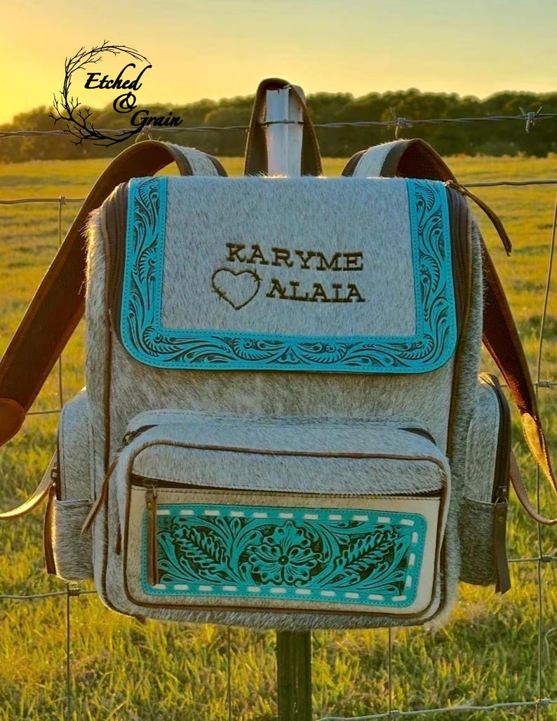 May include: A backpack made of grey and white cowhide featuring turquoise leather trim with embossed floral patterns. The front flap displays the text KARYME ALAIA above a heart outline. It includes a front zippered pocket and brown straps.