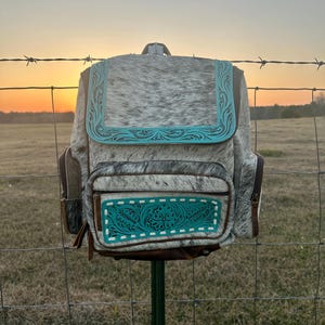 May include: A backpack crafted from grey and white cowhide leather featuring turquoise blue tooled leather accents on the front flap and the lower pocket. The bag includes brown leather side panels, a top handle, and visible zipper closures.