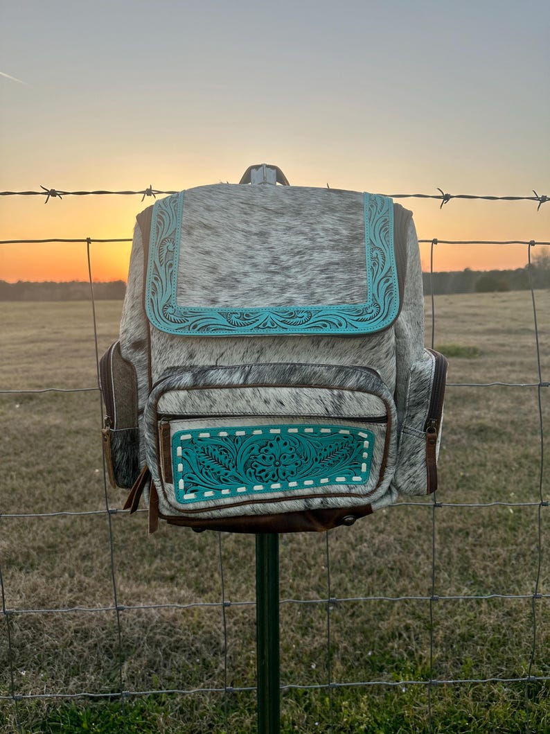 May include: A backpack crafted from grey and white cowhide leather featuring turquoise blue tooled leather accents on the front flap and the lower pocket. The bag includes brown leather side panels, a top handle, and visible zipper closures.