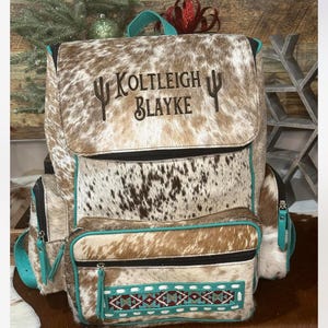 May include: A brown and white cowhide backpack featuring the embroidered text Koltleigh Blayke flanked by two cactus icons. The bag includes teal piping, a front pocket with a geometric beaded pattern, black zippers, and a top carry handle.