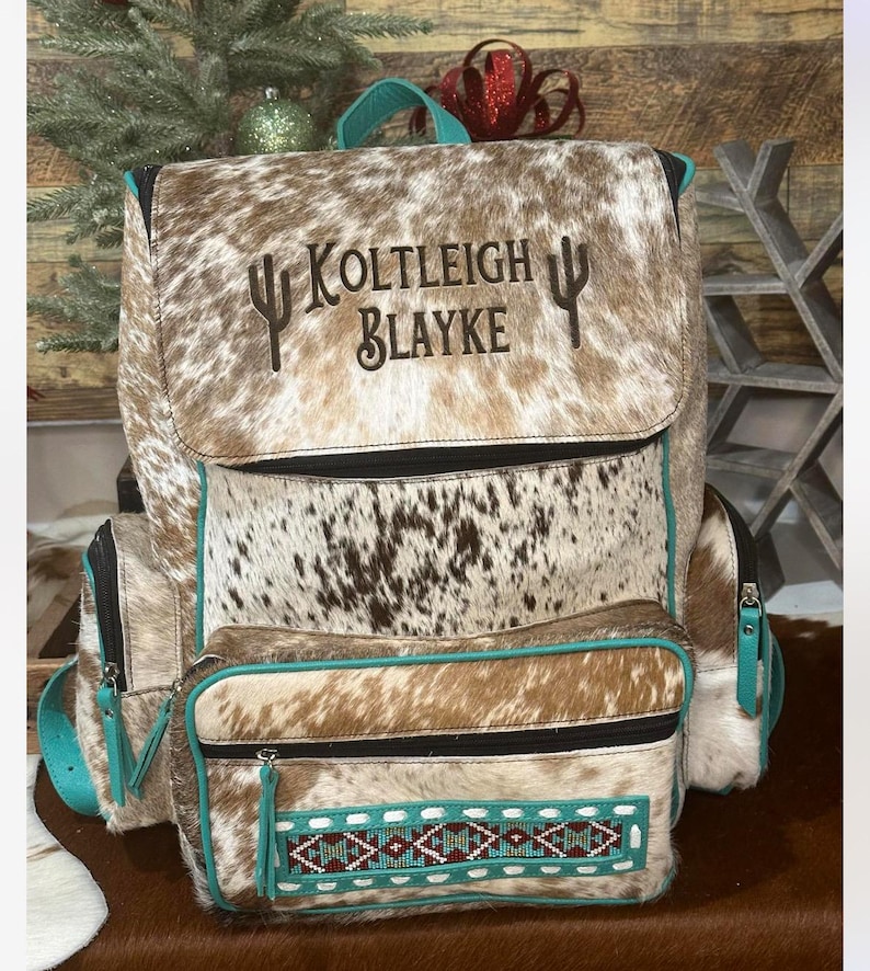 May include: A brown and white cowhide backpack featuring the embroidered text Koltleigh Blayke flanked by two cactus icons. The bag includes teal piping, a front pocket with a geometric beaded pattern, black zippers, and a top carry handle.