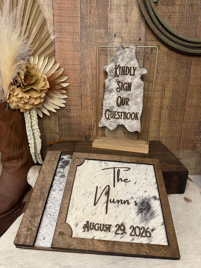 Peut inclure: Un livre d'or en bois avec une incrustation en peau de vache portant le texte "The Nunn's" et "AUGUST 29, 2026", accompagn&eacute; d'un panneau en peau de vache sur support en m&eacute;tal dor&eacute; indiquant "KINDLY SIGN OUR GUESTBOOK" avec des d&eacute;tails en bois.