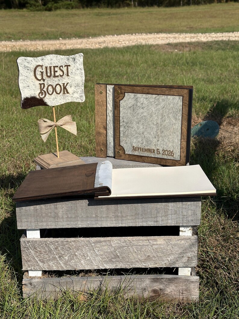 Peut inclure: Un panneau livre d'or avec une texture en peau de vache et le texte GUEST BOOK, accompagn&eacute; d'un livre d'or assorti avec une reliure en bois fonc&eacute;, une couverture en peau de vache et le texte grav&eacute; SEPTEMBER 5, 2026, et un livre ouvert.