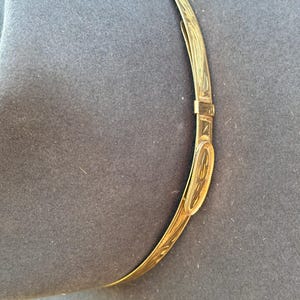 K&ouml;nnte beinhalten: Ein dunkelgrauer Filzhut mit einem d&uuml;nnen, goldfarbenen Hutband aus Metall. Das Band ist mit einem komplizierten, gravierten Blattmuster verziert und enth&auml;lt eine kleine, ovale Schnalle mit einem passenden strukturierten Design.