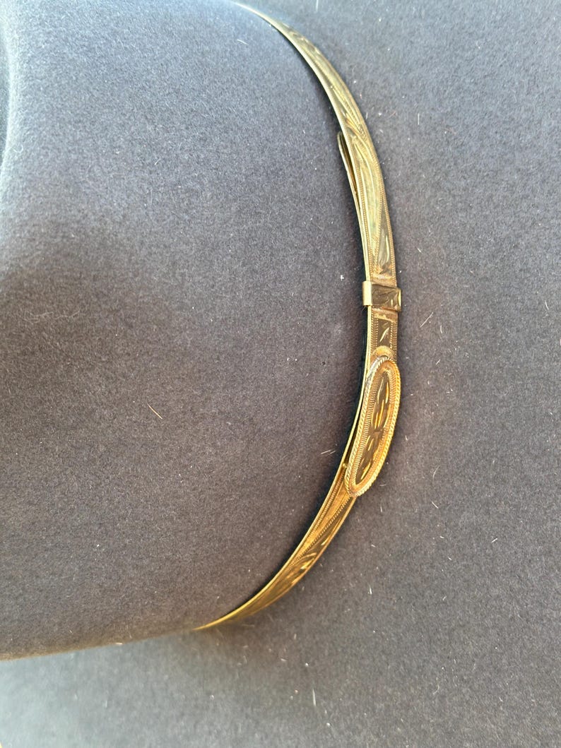 K&ouml;nnte beinhalten: Ein dunkelgrauer Filzhut mit einem d&uuml;nnen, goldfarbenen Hutband aus Metall. Das Band ist mit einem komplizierten, gravierten Blattmuster verziert und enth&auml;lt eine kleine, ovale Schnalle mit einem passenden strukturierten Design.