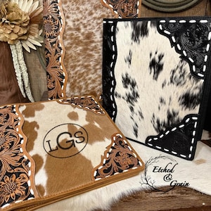 May include: Three rectangular binders featuring cowhide panels and dark leather corners with intricate floral tooling and white whipstitching. One binder displays the monogram LGS in a circular frame. The text Etched & Grain is visible.