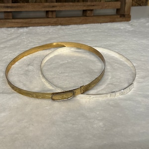 K&ouml;nnte beinhalten: Zwei d&uuml;nne, kreisf&ouml;rmige Metallarmreifen, einer in Goldoptik und einer in Silberoptik. Der goldene Armreif verf&uuml;gt &uuml;ber einen ovalen Verschluss mit eingraviertem Muster, w&auml;hrend beide B&auml;nder ein zartes, ge&auml;tztes Blumenmuster aufweisen.