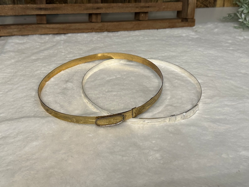 K&ouml;nnte beinhalten: Zwei d&uuml;nne, kreisf&ouml;rmige Metallarmreifen, einer in Goldoptik und einer in Silberoptik. Der goldene Armreif verf&uuml;gt &uuml;ber einen ovalen Verschluss mit eingraviertem Muster, w&auml;hrend beide B&auml;nder ein zartes, ge&auml;tztes Blumenmuster aufweisen.