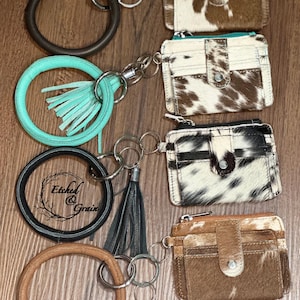 May include: Four keychains featuring circular wristlet rings in dark brown, turquoise, black, and tan. Each ring is attached to a matching leather tassel and a rectangular card holder wallet with a snap closure, showcasing various cowhide patterns.
