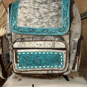 Op de afbeelding: Een rugzak met een zwart-wit koeienhuidpatroon en turquoise blauwe lederen accenten. De turquoise delen zijn voorzien van gedetailleerd bloemmotief in reli&euml;f en witte stiksels. Het heeft een klep, een voorvak met rits en zijvakken met bruine lederen afwerking.