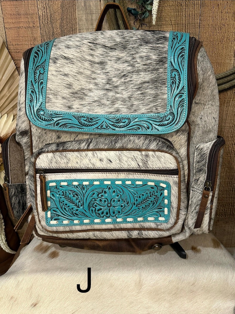 Op de afbeelding: Een rugzak met een zwart-wit koeienhuidpatroon en turquoise blauwe lederen accenten. De turquoise delen zijn voorzien van gedetailleerd bloemmotief in reli&euml;f en witte stiksels. Het heeft een klep, een voorvak met rits en zijvakken met bruine lederen afwerking.