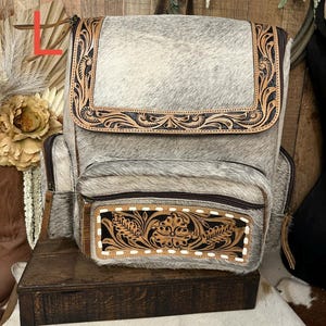 May include: A gray and white cowhide backpack featuring a top flap and front pocket with dark brown leather trim. The leather accents are decorated with intricate floral tooling and stitching. The bag includes side pockets and zipper closures.