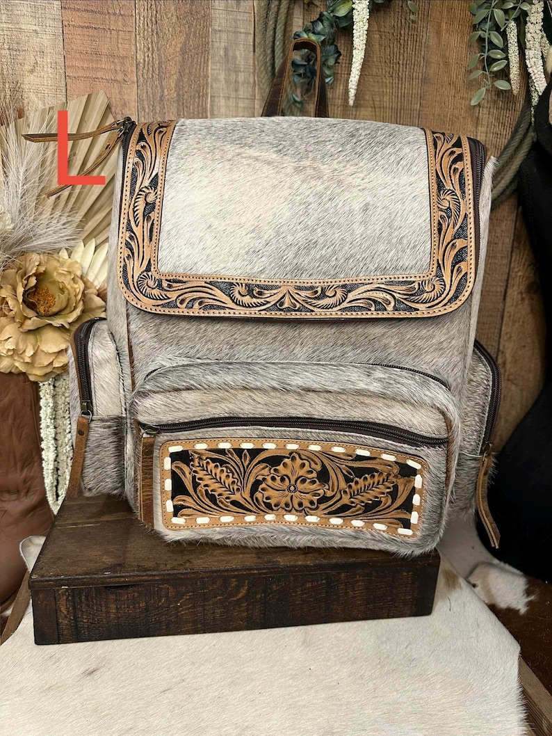 May include: A gray and white cowhide backpack featuring a top flap and front pocket with dark brown leather trim. The leather accents are decorated with intricate floral tooling and stitching. The bag includes side pockets and zipper closures.
