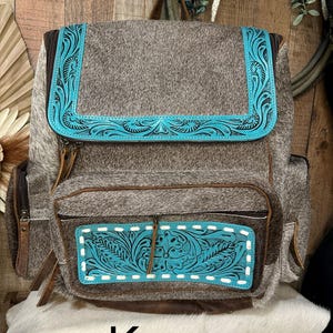 Op de afbeelding: Een rugzak van bruin en grijs koeienhuid met turquoise lederen accenten en gedetailleerd bloemmotief. De tas heeft een bovenflap, een voorvak met rits en witte stiksels, zijvakken en bruine lederen ritstrekkers.