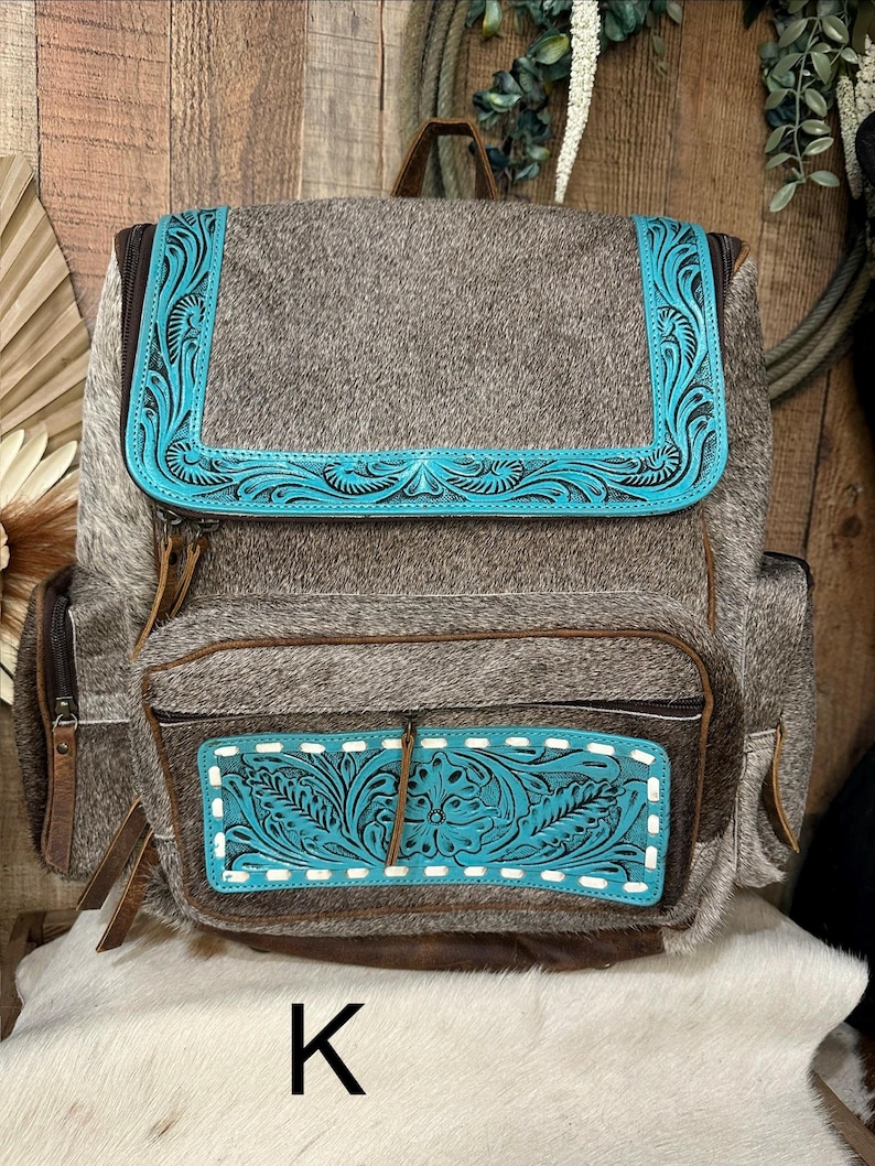 Op de afbeelding: Een rugzak van bruin en grijs koeienhuid met turquoise lederen accenten en gedetailleerd bloemmotief. De tas heeft een bovenflap, een voorvak met rits en witte stiksels, zijvakken en bruine lederen ritstrekkers.