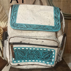 Op de afbeelding: Een rugzak van wit en zwart koeienhuid, geaccentueerd met turquoise lederen panelen met in reli&euml;f aangebrachte bloemmotieven. De tas heeft een bovenflap, een voorvak met rits en bruine lederen ritstrekkers. De tekst A1 is zichtbaar.