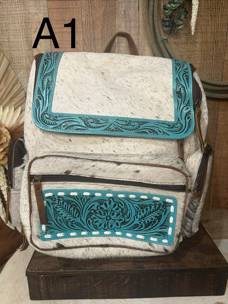 Op de afbeelding: Een rugzak van wit en zwart koeienhuid, geaccentueerd met turquoise lederen panelen met in reli&euml;f aangebrachte bloemmotieven. De tas heeft een bovenflap, een voorvak met rits en bruine lederen ritstrekkers. De tekst A1 is zichtbaar.