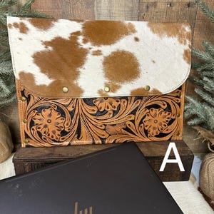May include: A laptop sleeve with a cowhide flap and a tooled leather base. The cowhide is brown and white, and the leather base features a floral design. The sleeve is closed with brass rivets and is sitting on a wooden surface.