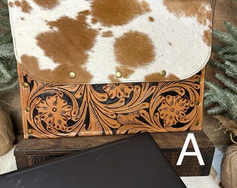 May include: A laptop sleeve with a cowhide flap and a tooled leather base. The cowhide is brown and white, and the leather base features a floral design. The sleeve is closed with brass rivets and is sitting on a wooden surface.
