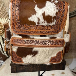 May include: A backpack crafted from brown and white cowhide featuring intricate floral tooling on the leather trim. The bag includes a large top flap, a zippered front pocket, and side compartments, all accented with detailed embossed leather borders.