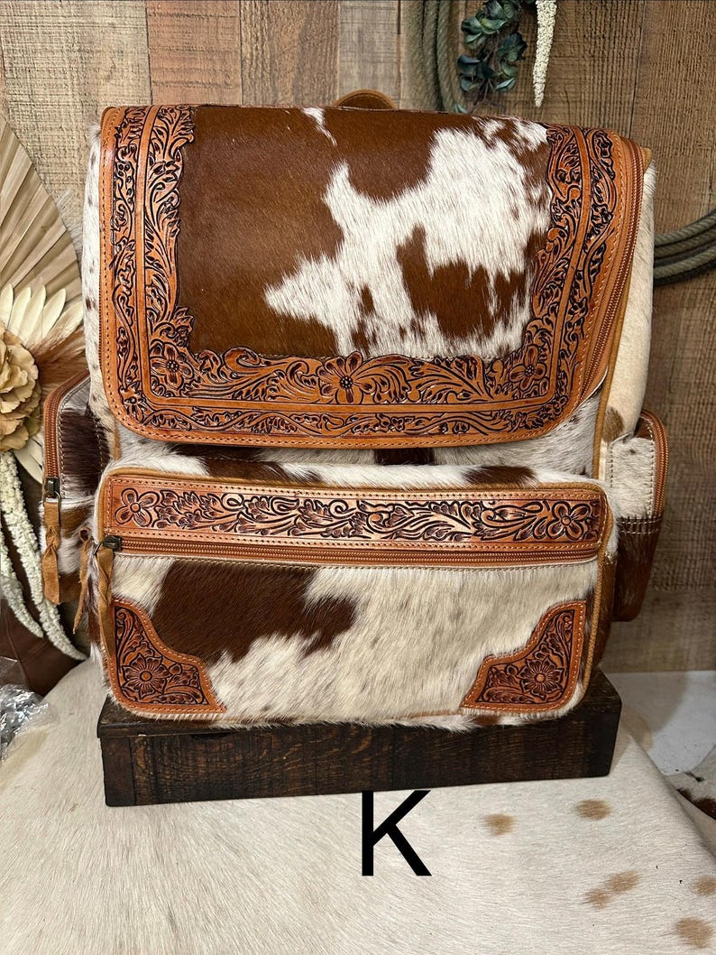 May include: A backpack crafted from brown and white cowhide featuring intricate floral tooling on the leather trim. The bag includes a large top flap, a zippered front pocket, and side compartments, all accented with detailed embossed leather borders.