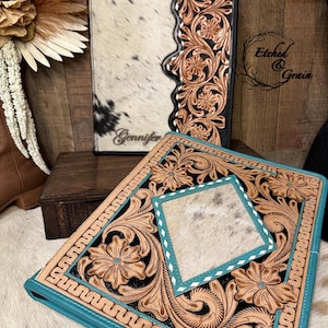 May include: Two rectangular leather portfolios featuring intricate floral tooling. The upright portfolio has white cowhide with black spots and the name Gennifer. The front portfolio has a central diamond-shaped white cowhide inlay with teal trim.