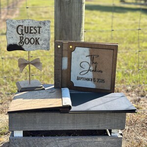 Peut inclure: Un livre d'or en bois avec une couverture en peau de vache portant l'inscription The Jenkins et SEPTEMBER 5 2026, accompagn&eacute; d'un panneau assorti sur pied indiquant GUEST BOOK, orn&eacute; d'un petit n&oelig;ud en toile de jute et d'un livre ouvert.