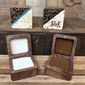May include: Two square wooden ring boxes with hinged lids. The lids feature a diagonal split design with dark brown embossed floral leather on one side and white hair-on hide on the other. One lid has a blue stripe, the other has the letters S&R.