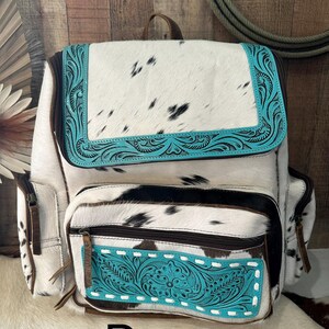 May include: A backpack crafted from white and black cowhide featuring a top flap and front pocket accented with bright turquoise leather panels. The turquoise sections are embossed with intricate floral patterns and white stitching details.