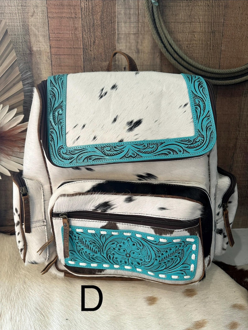 May include: A backpack crafted from white and black cowhide featuring a top flap and front pocket accented with bright turquoise leather panels. The turquoise sections are embossed with intricate floral patterns and white stitching details.