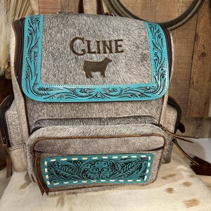 Op de afbeelding: Een grijs en wit koeienhuid rugzak met een turquoise leren klep voorzien van gedetailleerde bloemmotieven. Op de klep staat de tekst CLINE boven een silhouet van een koe. Een bijpassend turquoise leren paneel siert het voorvak.