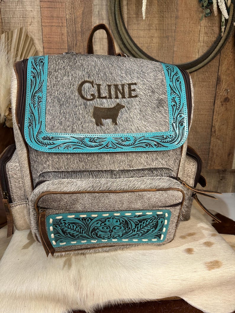Op de afbeelding: Een grijs en wit koeienhuid rugzak met een turquoise leren klep voorzien van gedetailleerde bloemmotieven. Op de klep staat de tekst CLINE boven een silhouet van een koe. Een bijpassend turquoise leren paneel siert het voorvak.