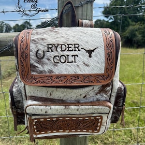 May include: A backpack made of brown and white cowhide featuring a top flap with tooled leather trim. The flap is engraved with the text RYDER COLT, a horseshoe icon, and a longhorn steer silhouette. A front pocket also displays tooled leather.