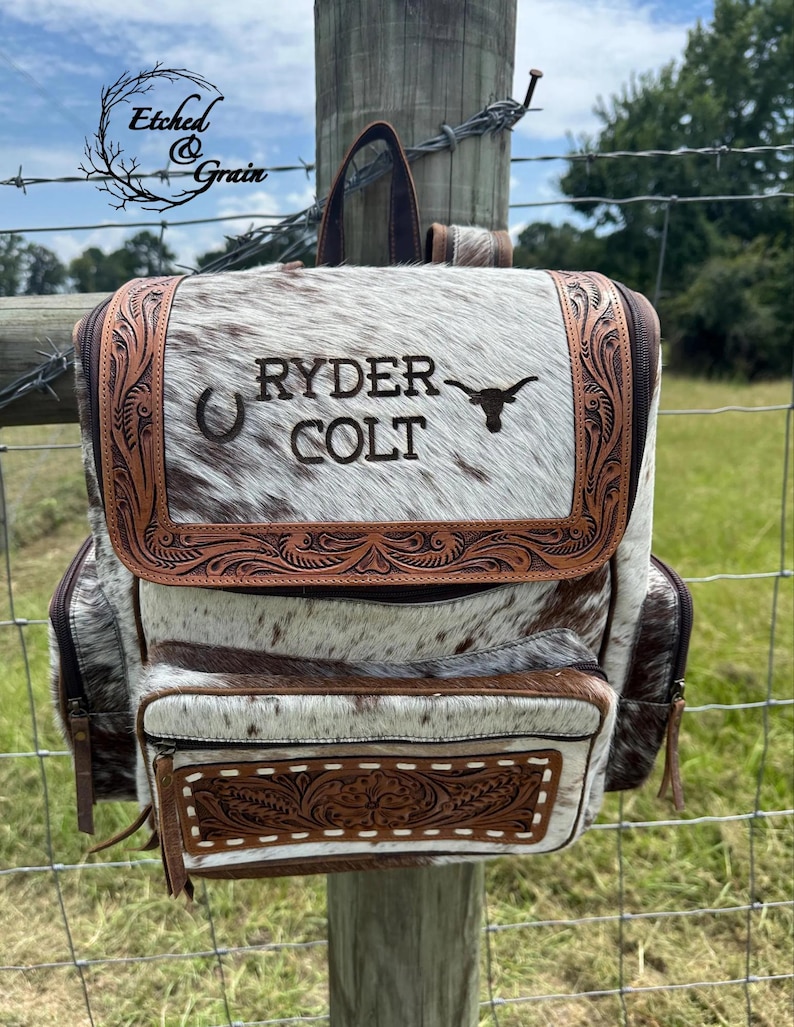 Op de afbeelding: Een rugzak van bruin en wit koeienhuid met een bovenflap met bewerkte lederen randen. De flap is gegraveerd met de tekst RYDER COLT, een hoefijzerpictogram en een silhouet van een longhorn-stier. Een voorvak is ook voorzien van bewerkt leer.