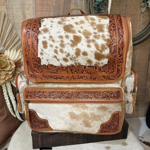 May include: A backpack featuring a brown and white cowhide pattern with tan leather trim. The leather sections are decorated with intricate floral tooling. It includes a large top flap, a front zippered pocket, and side pockets with zippers.