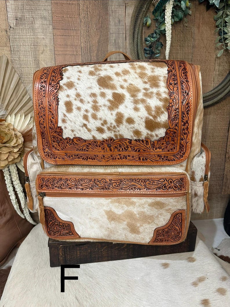 May include: A backpack featuring a brown and white cowhide pattern with tan leather trim. The leather sections are decorated with intricate floral tooling. It includes a large top flap, a front zippered pocket, and side pockets with zippers.