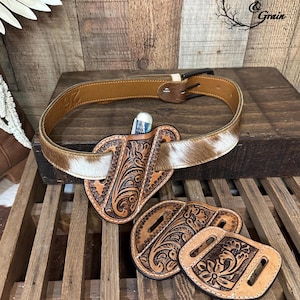 May include: A brown leather belt with a cowhide pattern, featuring a floral-tooled leather knife sheath threaded onto the strap. Two additional floral-tooled leather belt slides are positioned below. The text Etched & Grain is visible.