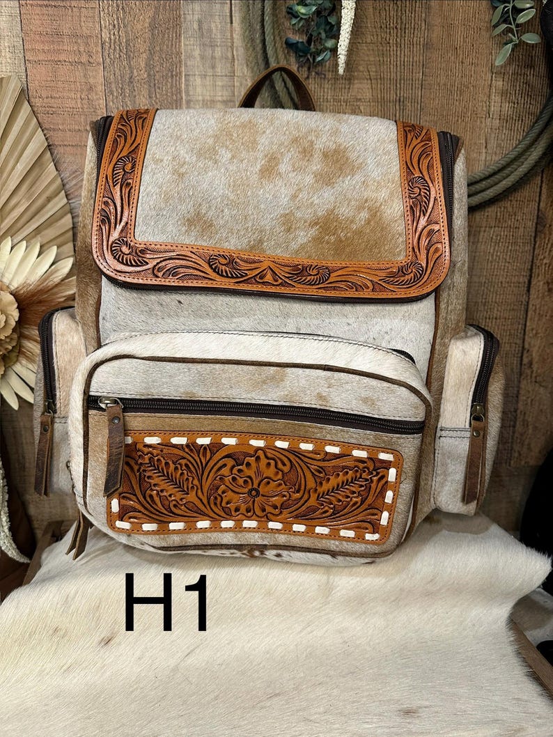 Op de afbeelding: Een rugzak van beige en witte koeienhuid met een bovenflap en voorvak met bruine lederen afwerking. De lederen delen zijn versierd met bloemmotieven en witte stiksels. De tekst H1 is hieronder zichtbaar.