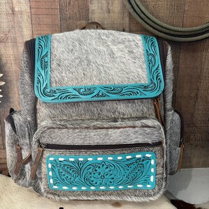 May include: A backpack crafted from grey and white cowhide featuring turquoise leather accents with intricate floral tooling. The bag includes a top flap, a front zippered pocket, and side pockets, all secured with brown zippers and leather pulls.