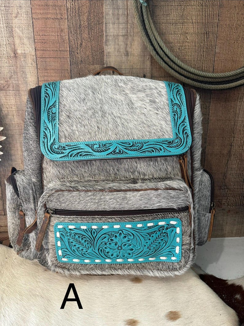 May include: A backpack crafted from grey and white cowhide featuring turquoise leather accents with intricate floral tooling. The bag includes a top flap, a front zippered pocket, and side pockets, all secured with brown zippers and leather pulls.