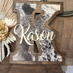 Peut inclure: Une grande lettre K en bois avec un cadre teinté brun foncé et une incrustation à motif de peau de vache noire et blanche. Le nom Kason est écrit dans une police cursive en bois de couleur claire et superposé au centre de la lettre K.