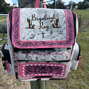May include: A backpack made of white and black cowhide with pink leather trim. The front flap features the engraved text "Bryleigh Ray" flanked by two cactus icons and a cross. The front pocket has a floral embossed pink leather panel.