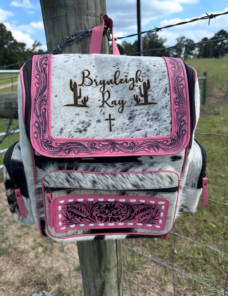May include: A backpack made of white and black cowhide with pink leather trim. The front flap features the engraved text "Bryleigh Ray" flanked by two cactus icons and a cross. The front pocket has a floral embossed pink leather panel.