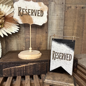 May include: Two "Reserved" signs. One is a cowhide-patterned sign on a wooden stand. The other is a cowhide-patterned banner with a wooden base. The signs have a rustic, western theme.